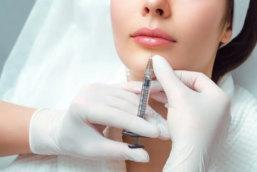 "Close-up of a woman's face receiving a dermal filler injection below the lips from a gloved professional, highlighting the treatment area around the lower lip and chin.