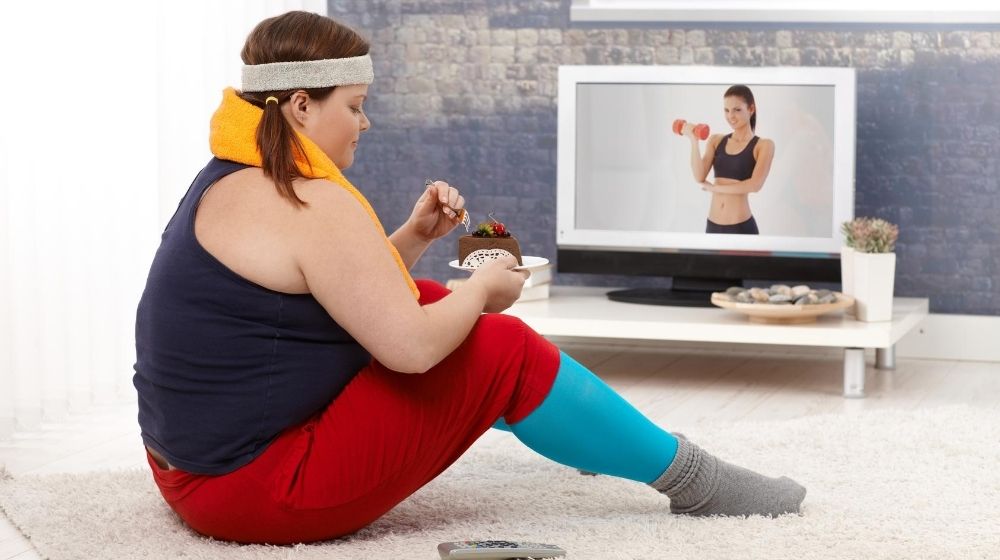 an overweight girl wearing sport cloth and eating cake while watching fitness show | Feature | Why Can't I Lose Weight!? | 6 Stubborn Weight Loss FAQs